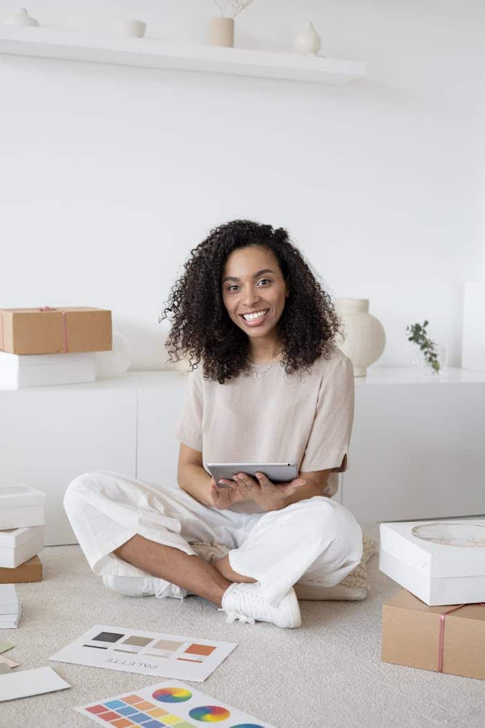A confident young woman sitting among packages and papers while using a tablet, showcasing modern entrepreneurship.